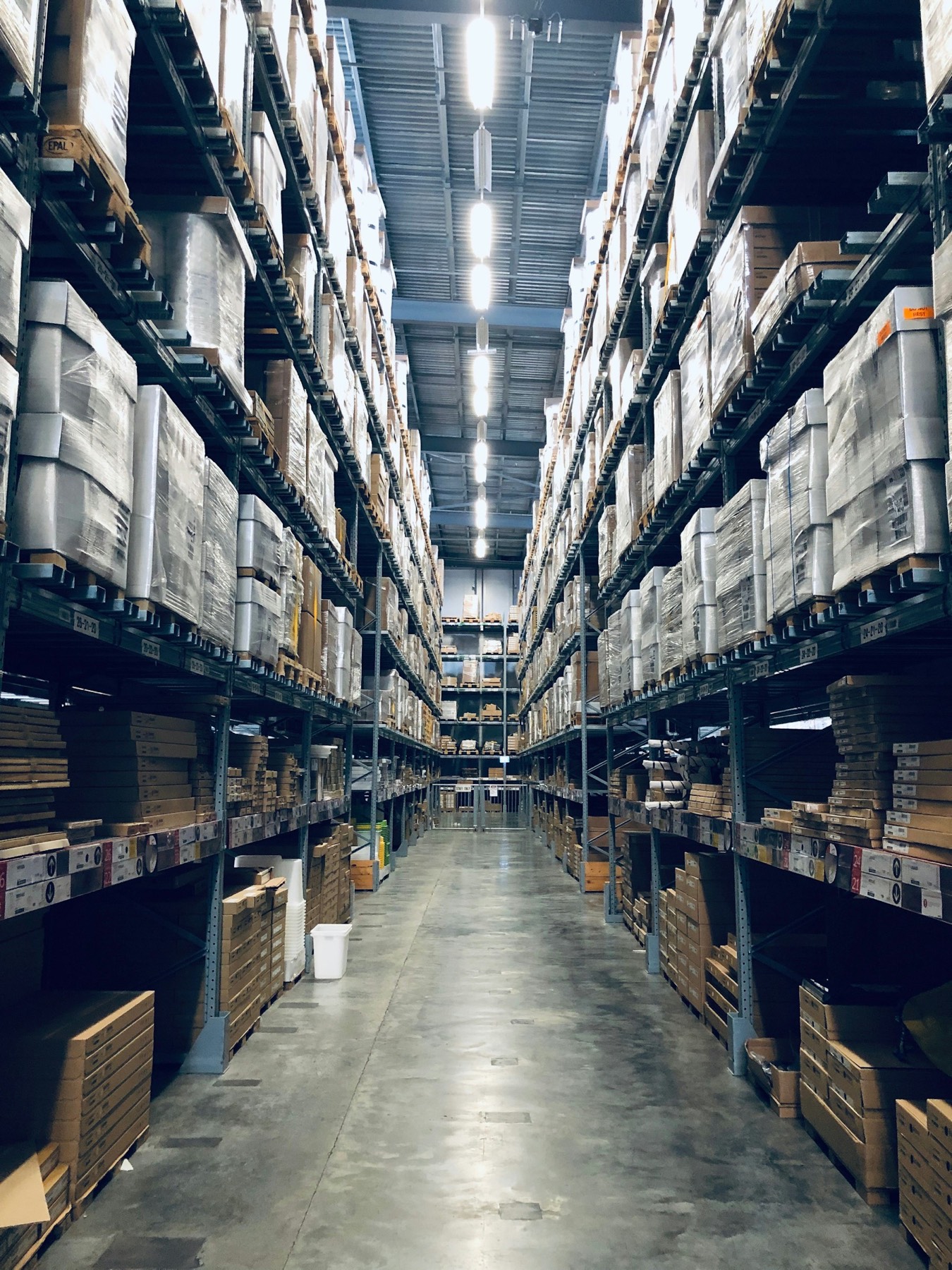 Warehouse aisle between tall pallet racking, shrink-wrapped pallets on both sides, concrete floor leading to the far wall